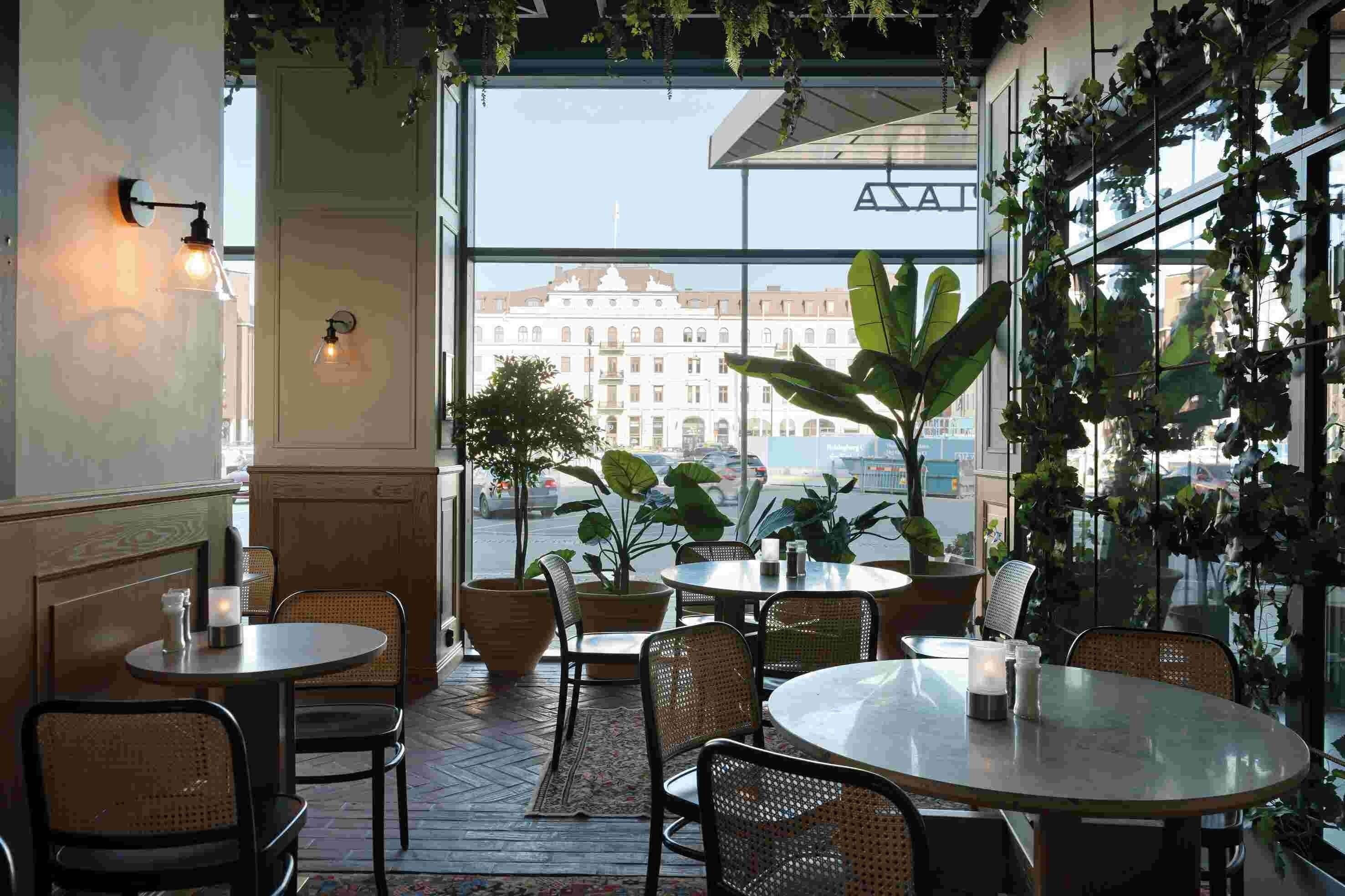 An indoor dining area with round tables and wicker chairs, large potted plants, and a view of a historic building through large windows.