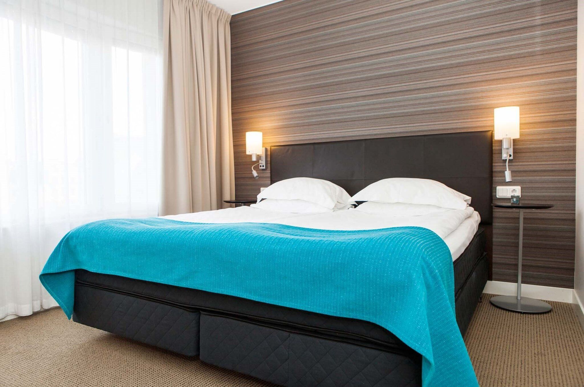 A hotel room with a bed, two bedside lamps, a nightstand, and a window with curtains.