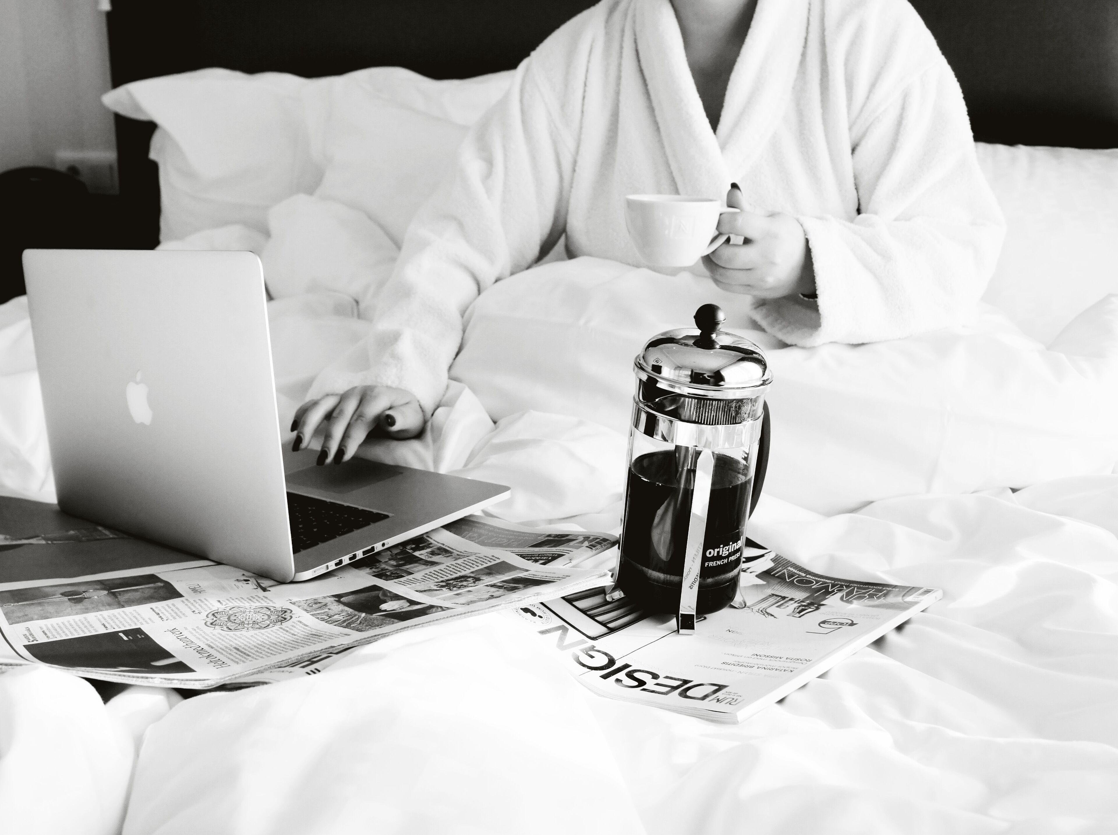A person in a white robe using a laptop on a bed with a newspaper, a coffee press, and a cup.