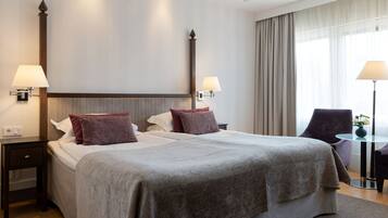 Habitación de hotel con una cama grande, dos mesitas de noche con lámparas, una silla y una pequeña mesa con un florero.