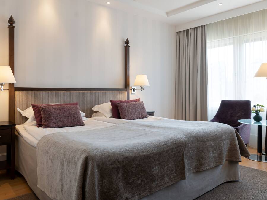 Habitación de hotel con una cama grande, dos mesitas de noche con lámparas, una silla y una pequeña mesa con un florero.
