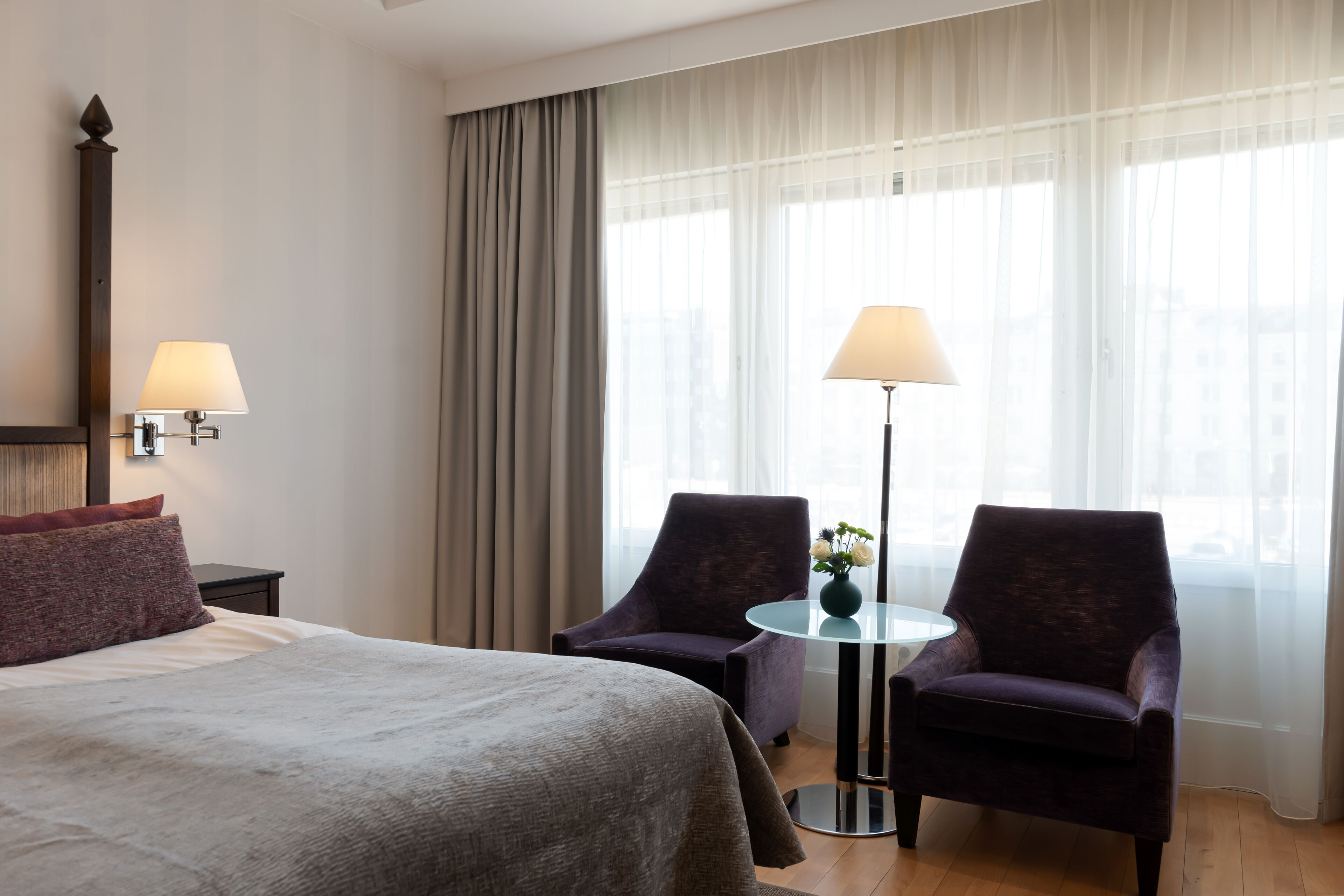Una habitación de hotel con una cama grande, dos sillones, una mesita con un florero y una lámpara.