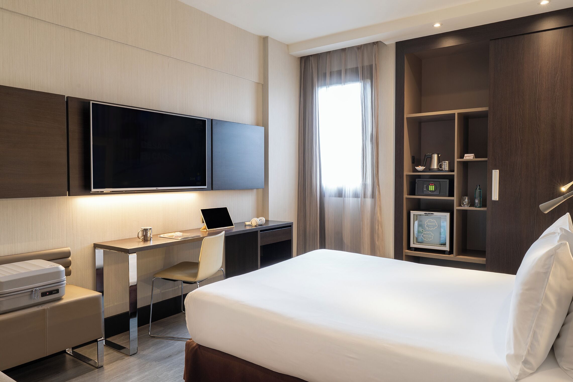 Quarto de hotel com cama, escrivaninha, cadeira, televisão e estantes embutidas com diversos objetos.