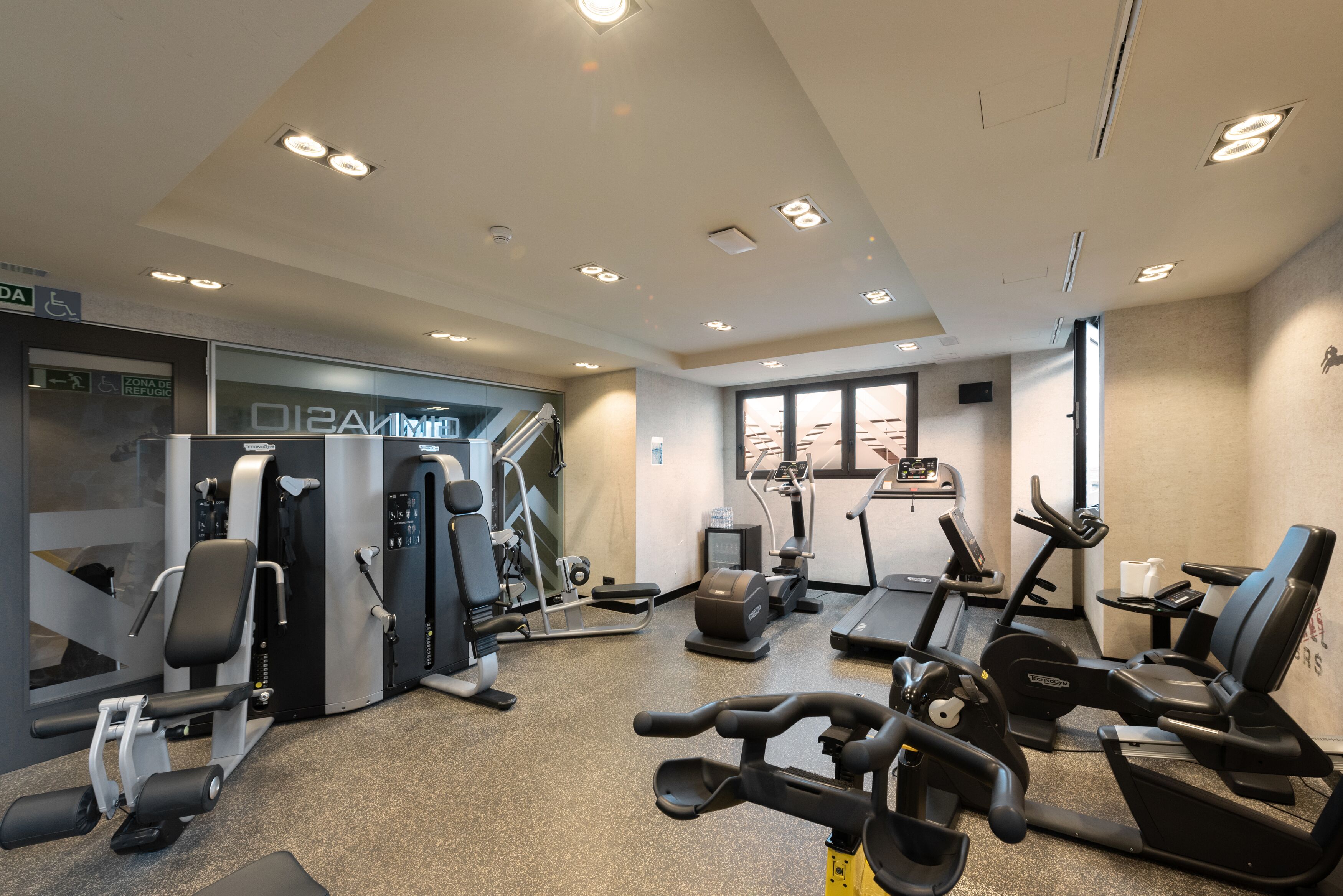 A gym with various exercise equipment, including treadmills and weight machines, and a small window providing natural light.