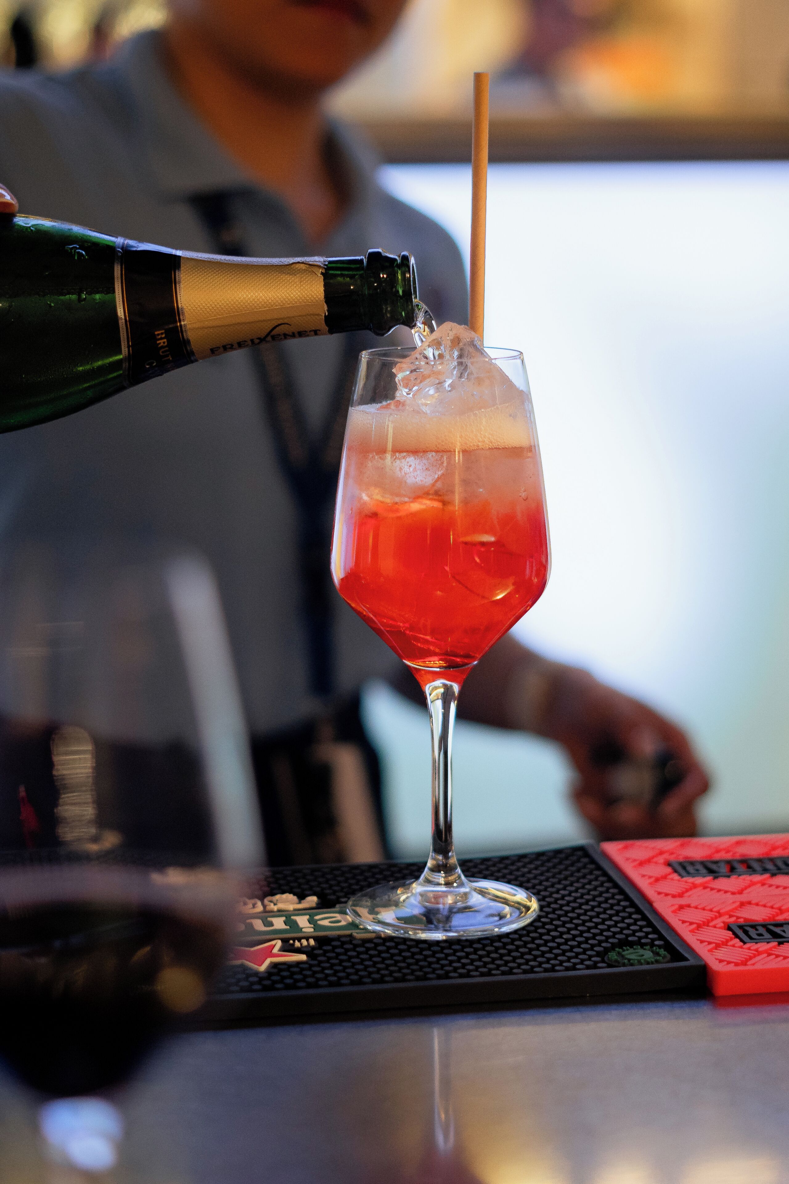 A bar setting with a glass being filled with a red liquid, a bottle of champagne, and a bar mat.