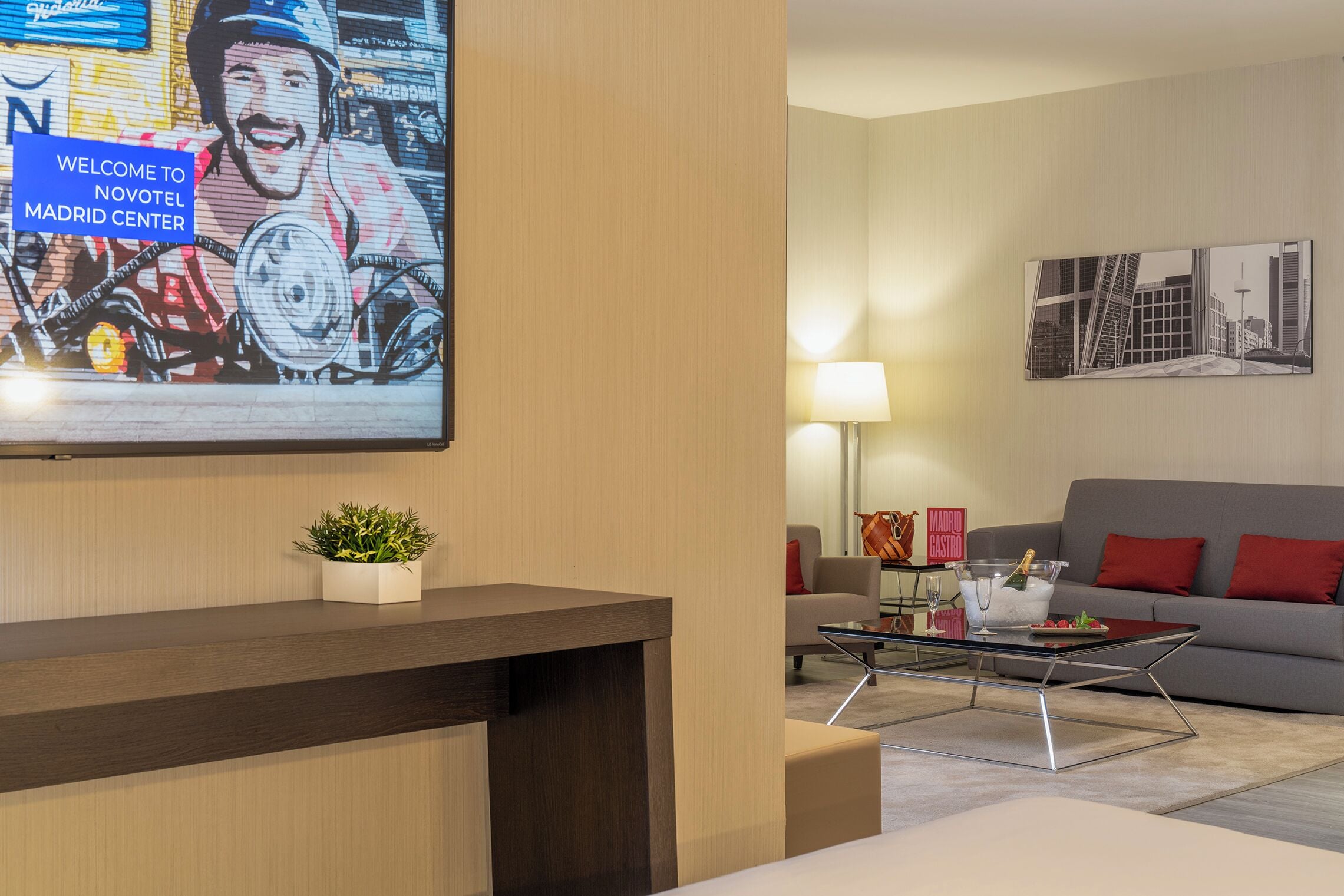 Een moderne hotel lobby met een receptiebalie, een bank, een salontafel en een aan de muur bevestigde televisie waarop een welkomstboodschap staat.