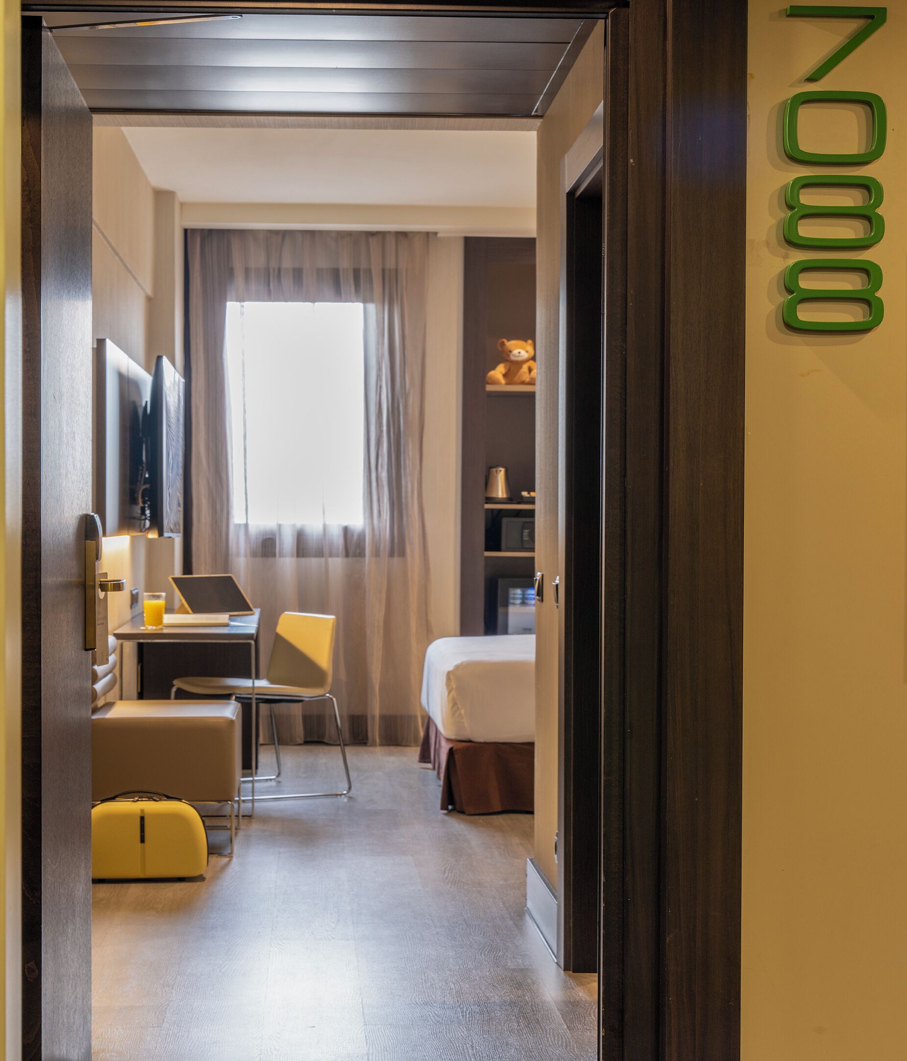 Habitación de hotel con cama, escritorio con silla, mesita de noche con lámpara, maleta y ventana con cortinas.
