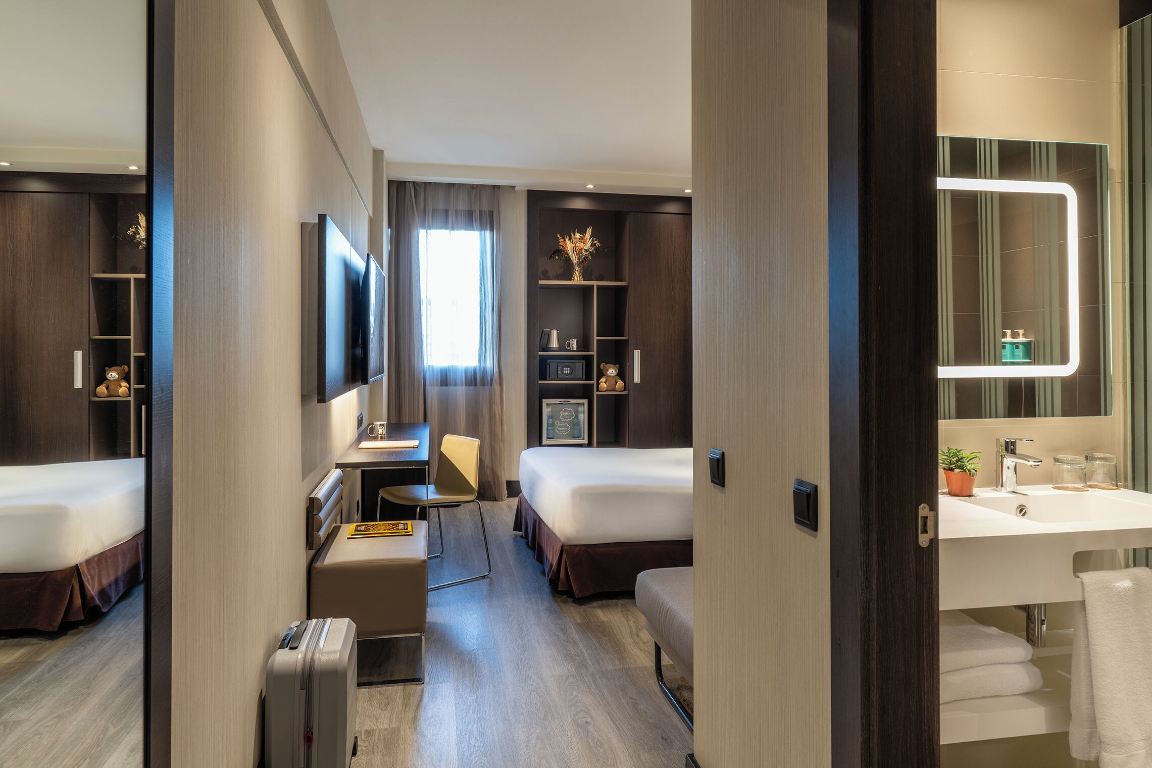 A modern hotel room with a bed, desk, chair, and a bathroom with a sink and mirror.