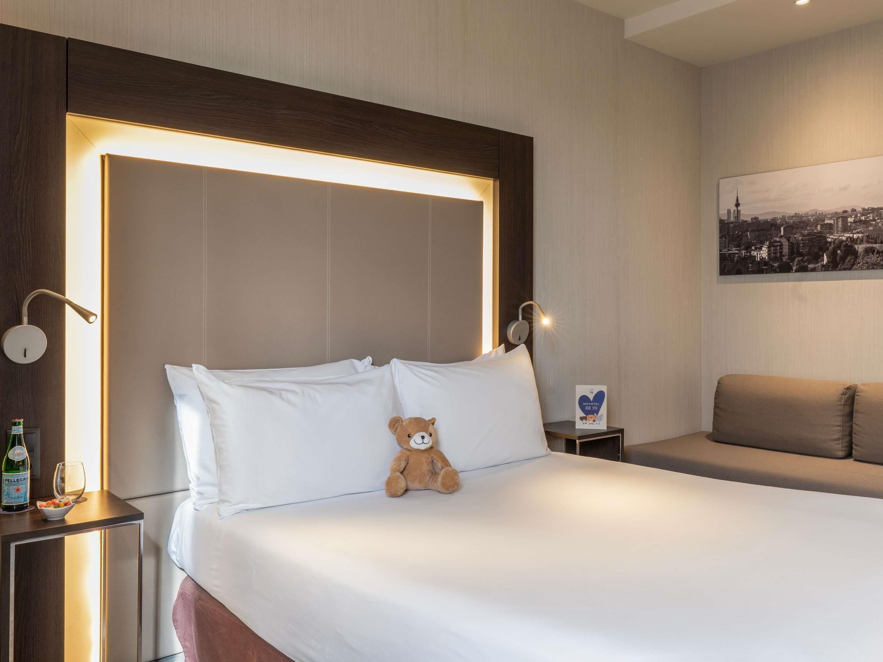 Habitación de hotel con una cama grande, cabecera con iluminación integrada, mesita de noche con una botella y un vaso, un osito de peluche en la cama y un cuadro de un paisaje urbano en la pared.