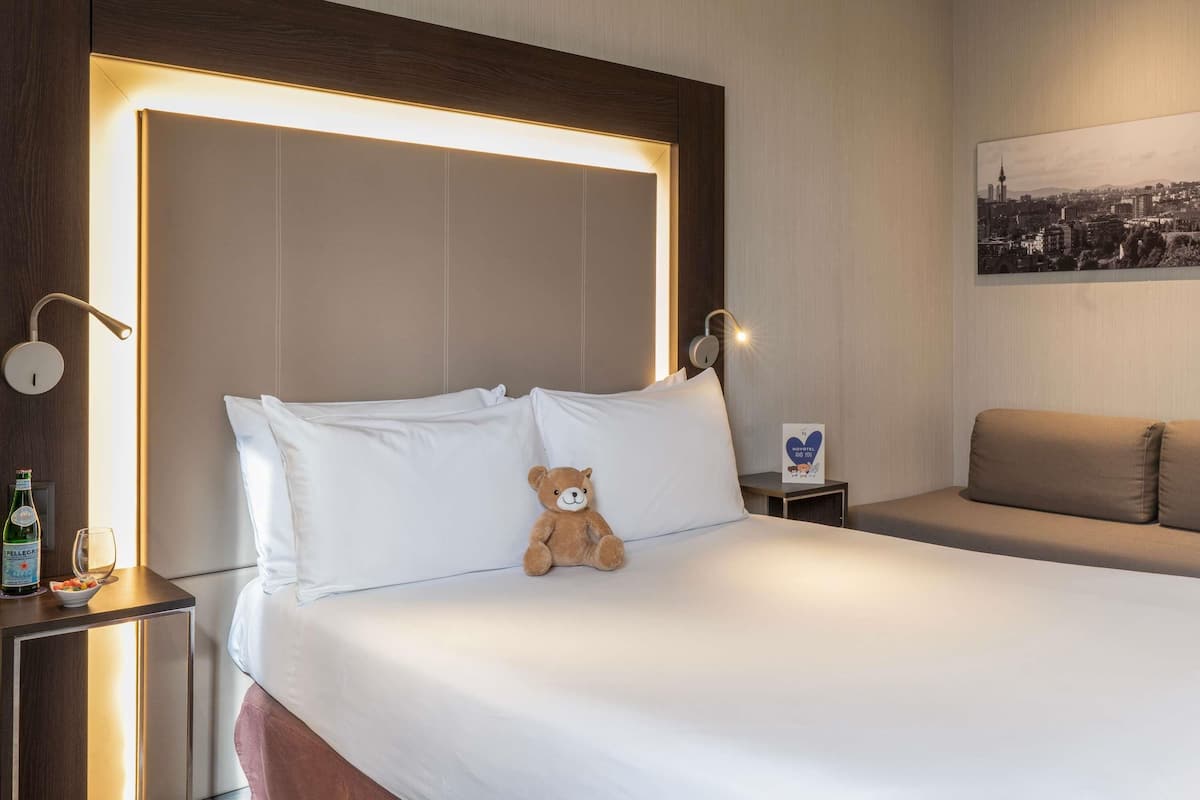 A hotel room with a large bed, a headboard with integrated lighting, a bedside table with a bottle and glass, a teddy bear on the bed, and a cityscape painting on the wall.