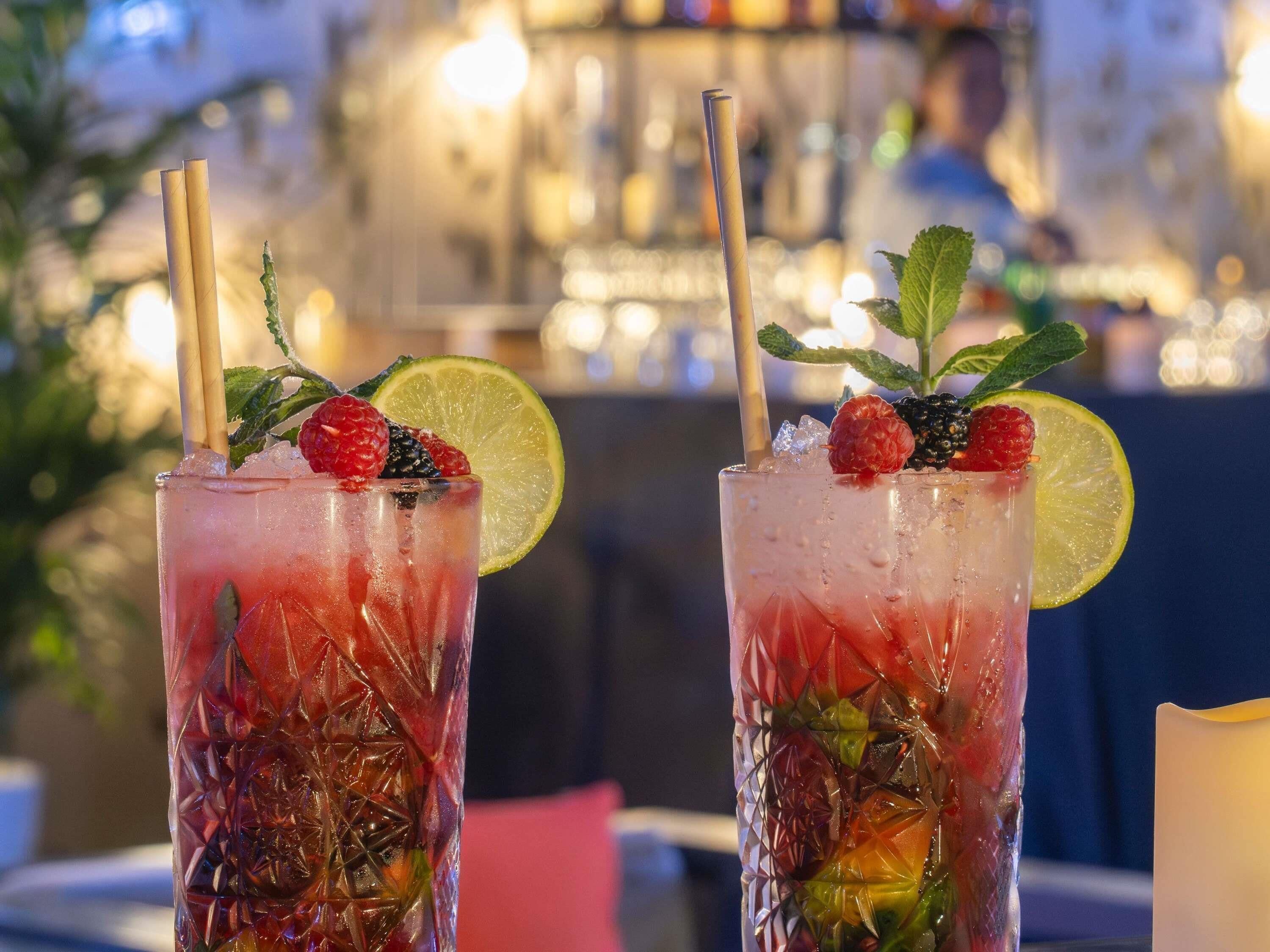 Two tall glasses of a red beverage with ice, garnished with raspberries, mint, and a slice of lime, with straws.