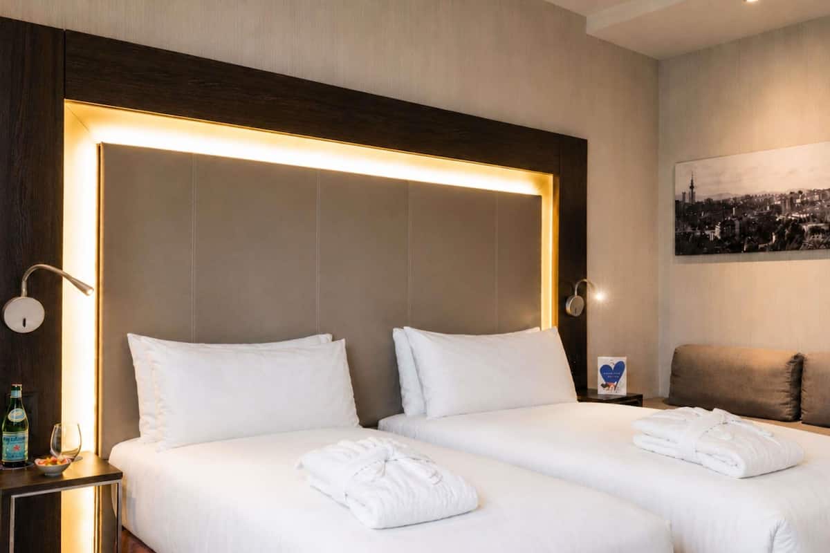 A hotel room with two beds, a headboard with integrated lighting, a bedside table with a bottle and glass, and a cityscape painting on the wall.