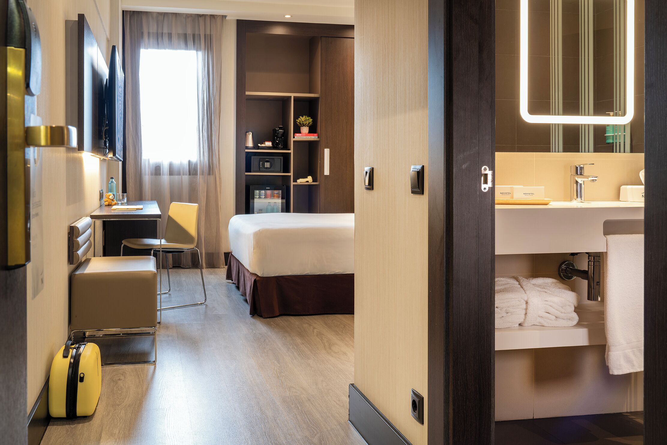 A hotel room with a bed, a desk, a chair, a suitcase, a minibar, and a bathroom with a mirror and towel rack.