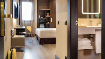 A hotel room with a bed, a desk, a chair, a suitcase, a minibar, and a bathroom with a mirror and towel rack.