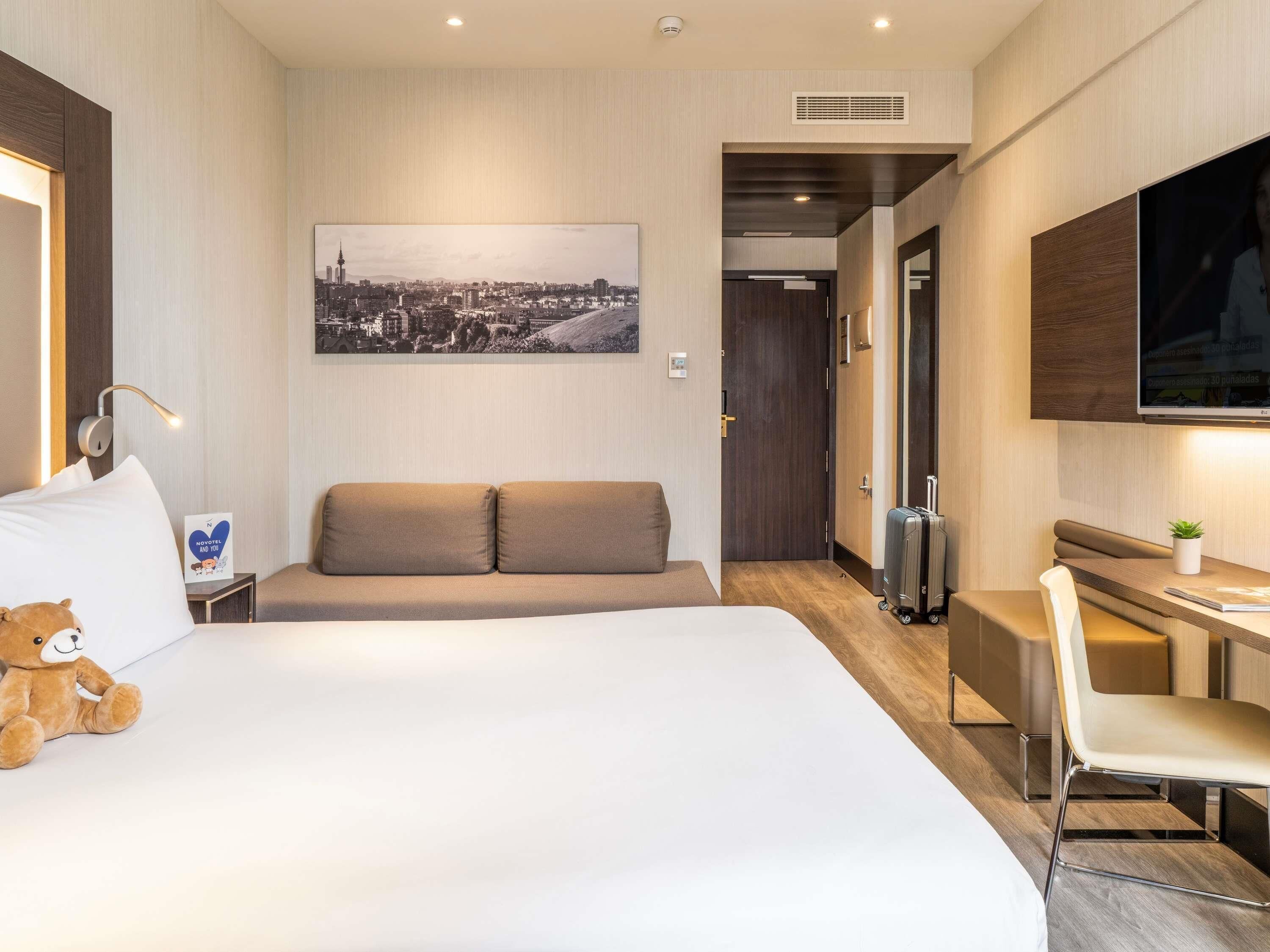 A hotel room with a bed, a desk, a chair, a sofa, a television, a window, and a cityscape view.