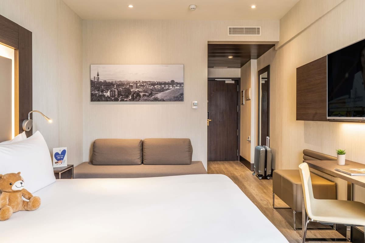 Ein Hotelzimmer mit Bett, Schreibtisch, Stuhl, Sofa, Fernseher und einem Fenster mit Blick auf die Stadt.