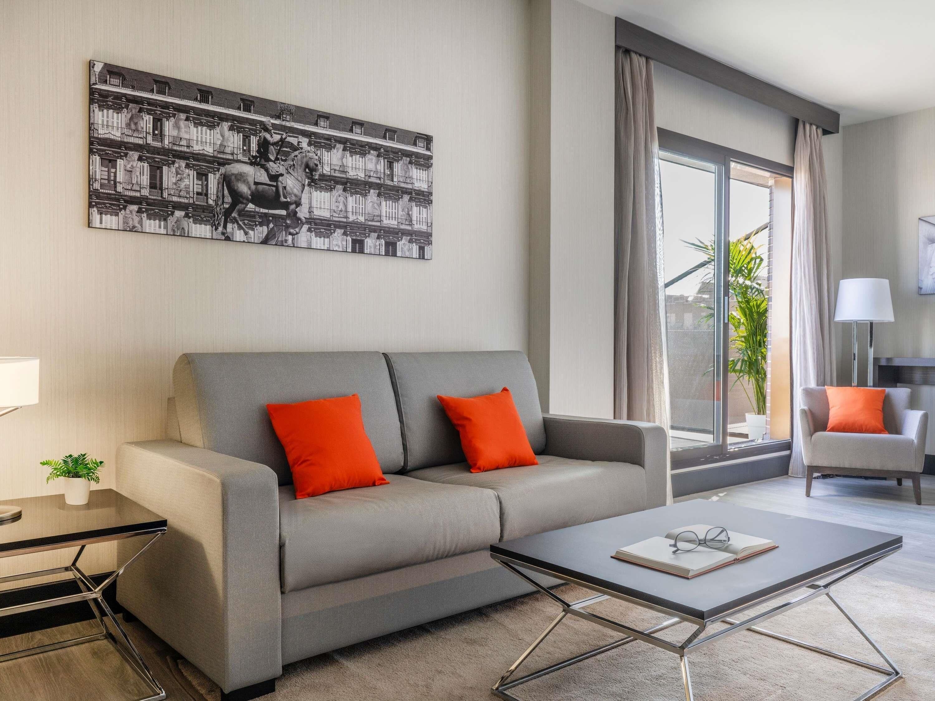 A modern living room with a grey sofa, two orange cushions, a glass coffee table, and a large window with curtains.