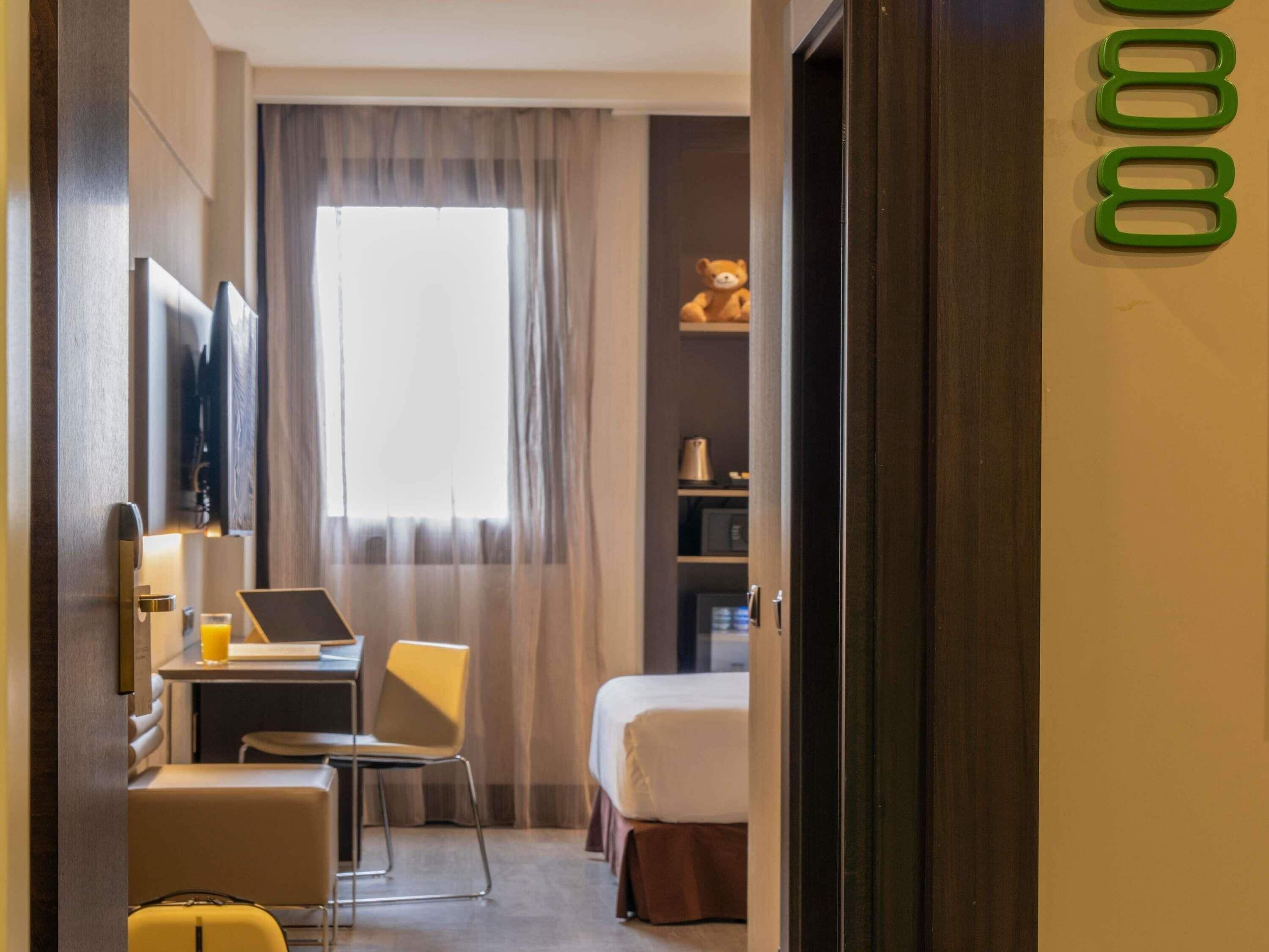 Habitación de hotel con cama, escritorio con una computadora portátil, silla, maleta y ventana con cortinas.