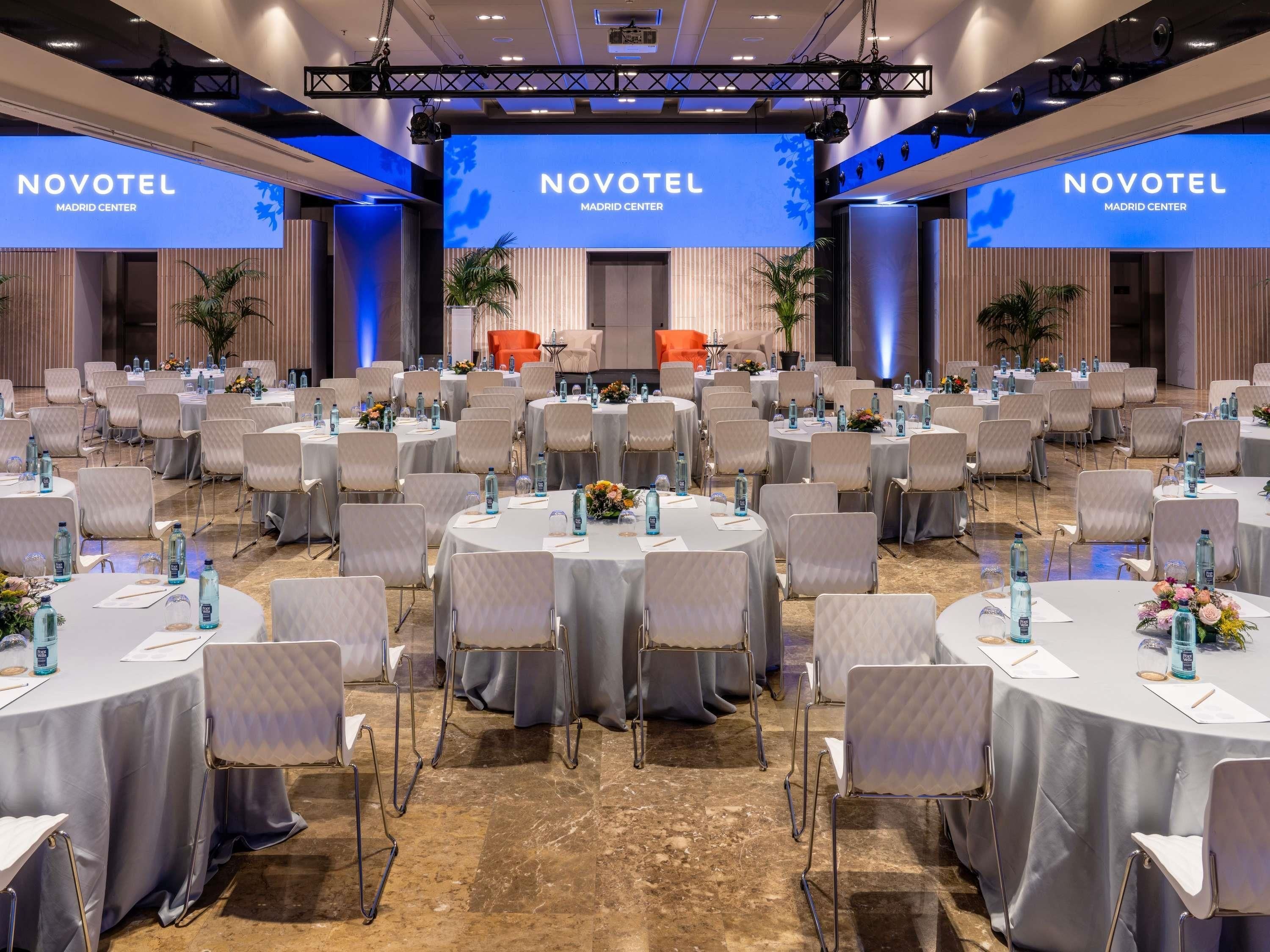 Salón de conferencias del Novotel Madrid Center con mesas redondas, sillas y un escenario con una pantalla grande.