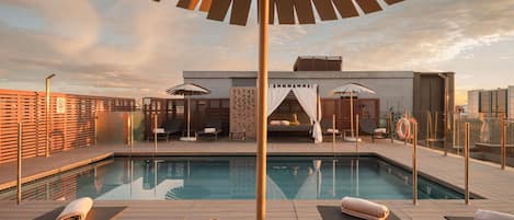 Un área de piscina en la azotea con sillones de descanso, una mesa con una taza de café y sombrillas.
