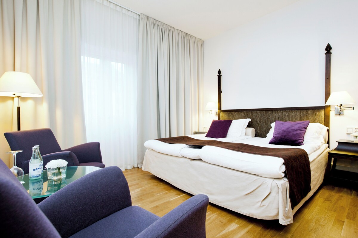 A hotel room with a bed, a glass table with a bottle, a chair, and a lamp.