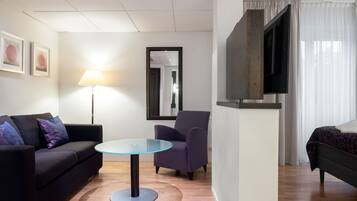 A modern hotel room with a sofa, armchair, round glass table, and a bed.