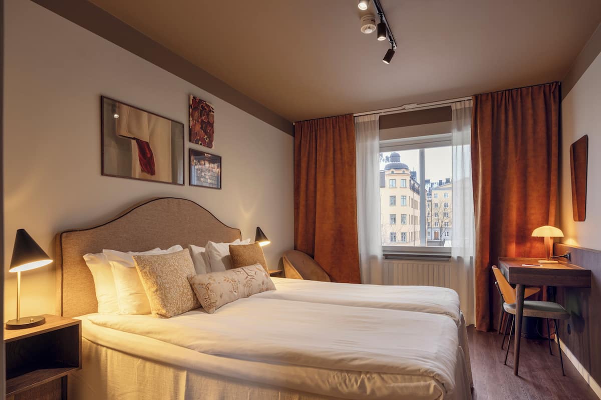 A hotel room with a large bed, bedside tables, a desk with a chair, and a window with curtains.