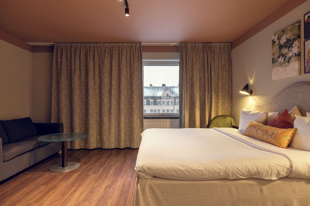 A hotel room with a bed, a sofa, a small table, and a window with curtains.