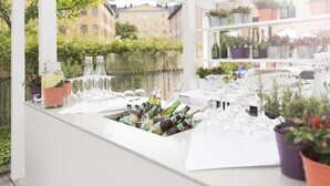 Zona de bar con botellas y vasos, plantas en macetas y vistas a edificios al fondo.