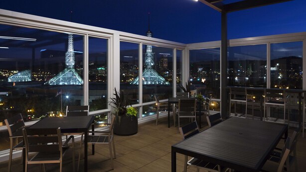 Una terraza en la azotea con mesas y sillas, amplias ventanas que ofrecen vistas a un paisaje urbano y estructuras iluminadas.
