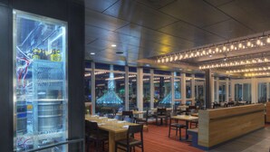 Un restaurante con zona de bar, mesas, sillas y amplios ventanales con vistas a la ciudad.