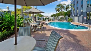 A pool area with lounge chairs and umbrellas, surrounded by palm trees and a building.