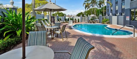 A pool area with lounge chairs and umbrellas, surrounded by palm trees and a building.