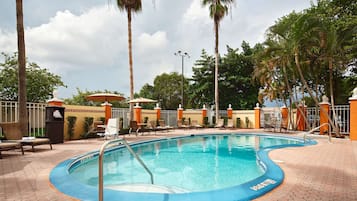 Une piscine entourée de palmiers et d’une clôture, avec des chaises longues et des parasols.