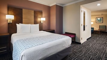Une chambre d’hôtel avec un grand lit, deux lampes de chevet, un petit banc, une porte donnant sur une autre pièce et un sol recouvert de moquette.