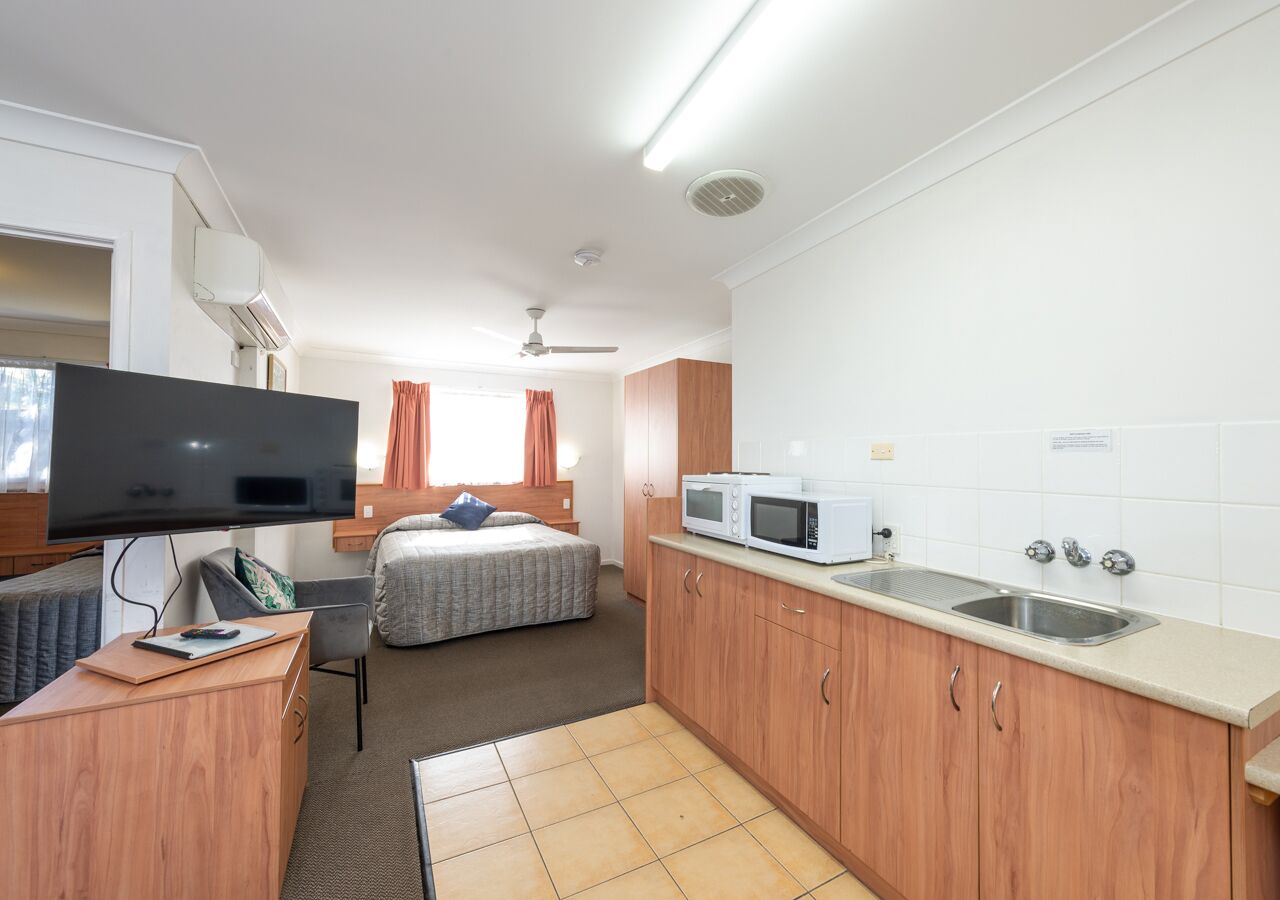 A compact hotel room with a bed, a desk with a computer, a TV, a kitchenette with a microwave and sink, and a ceiling fan.