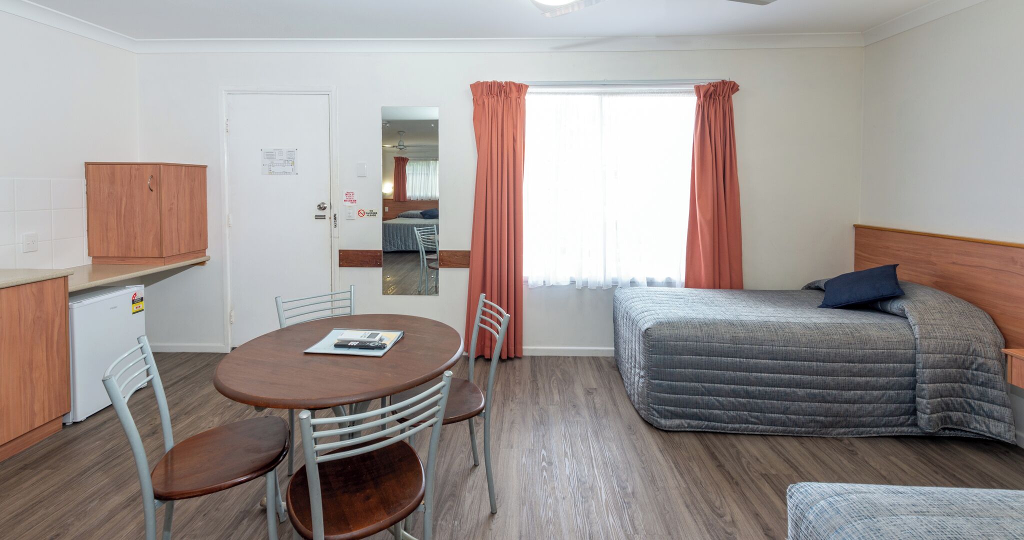Quarto compacto com cama, uma pequena mesa com cadeiras, cozinha compacta e um espelho.