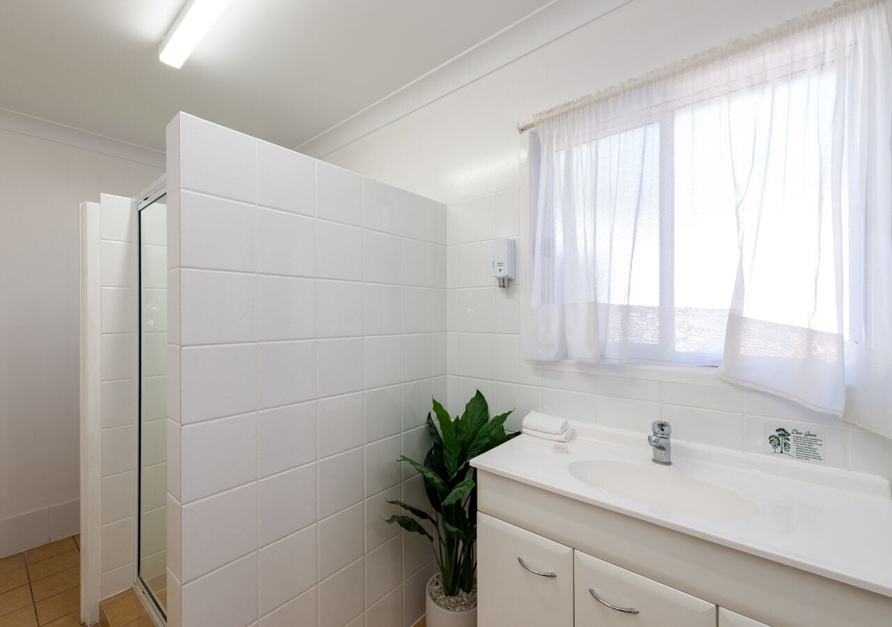 Ein Badezimmer mit weißen Fliesen, einer Duschkabine aus Glas, einem Waschbecken mit Spiegel und einem Fenster mit transparenten Vorhängen.