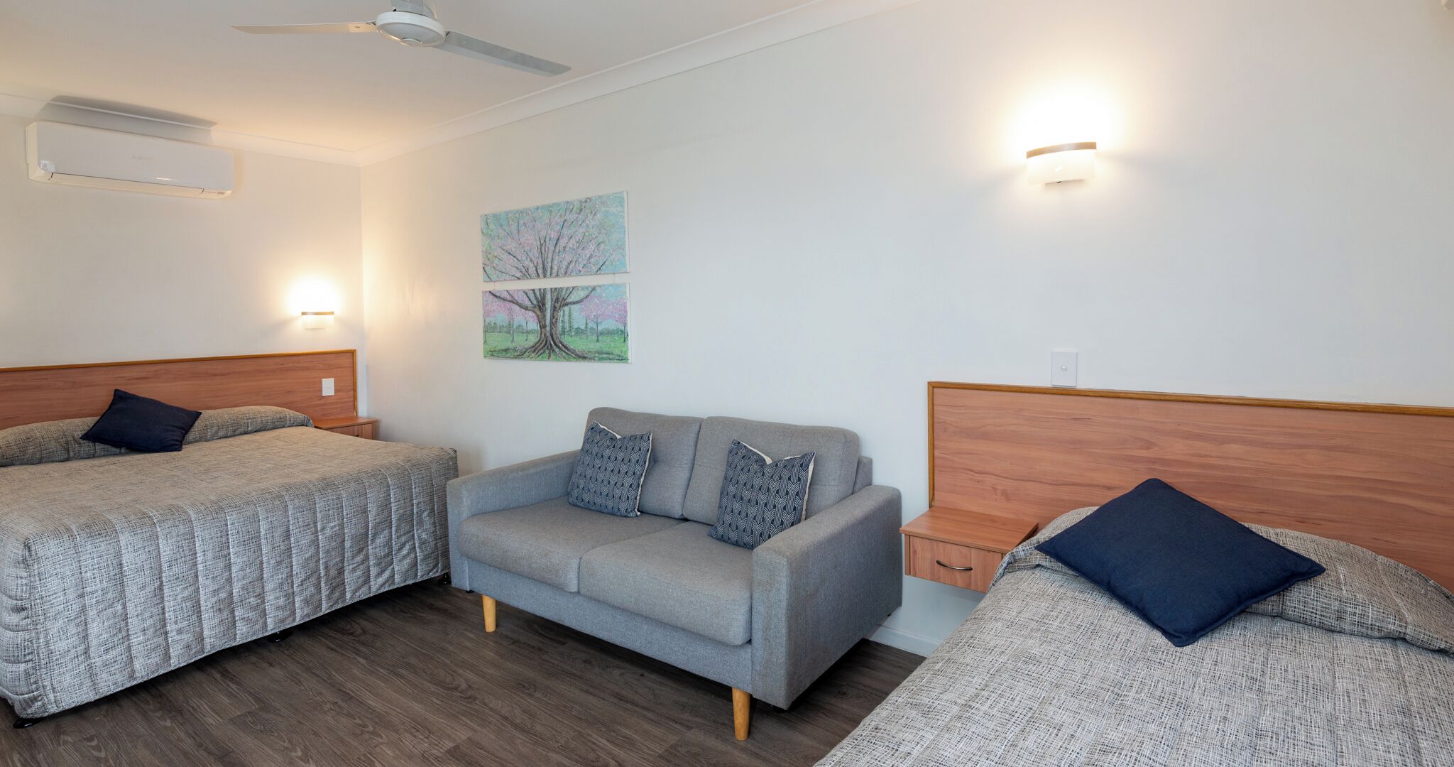 A hotel room with two single beds, a sofa, a ceiling fan, wall-mounted lights, and a painting.