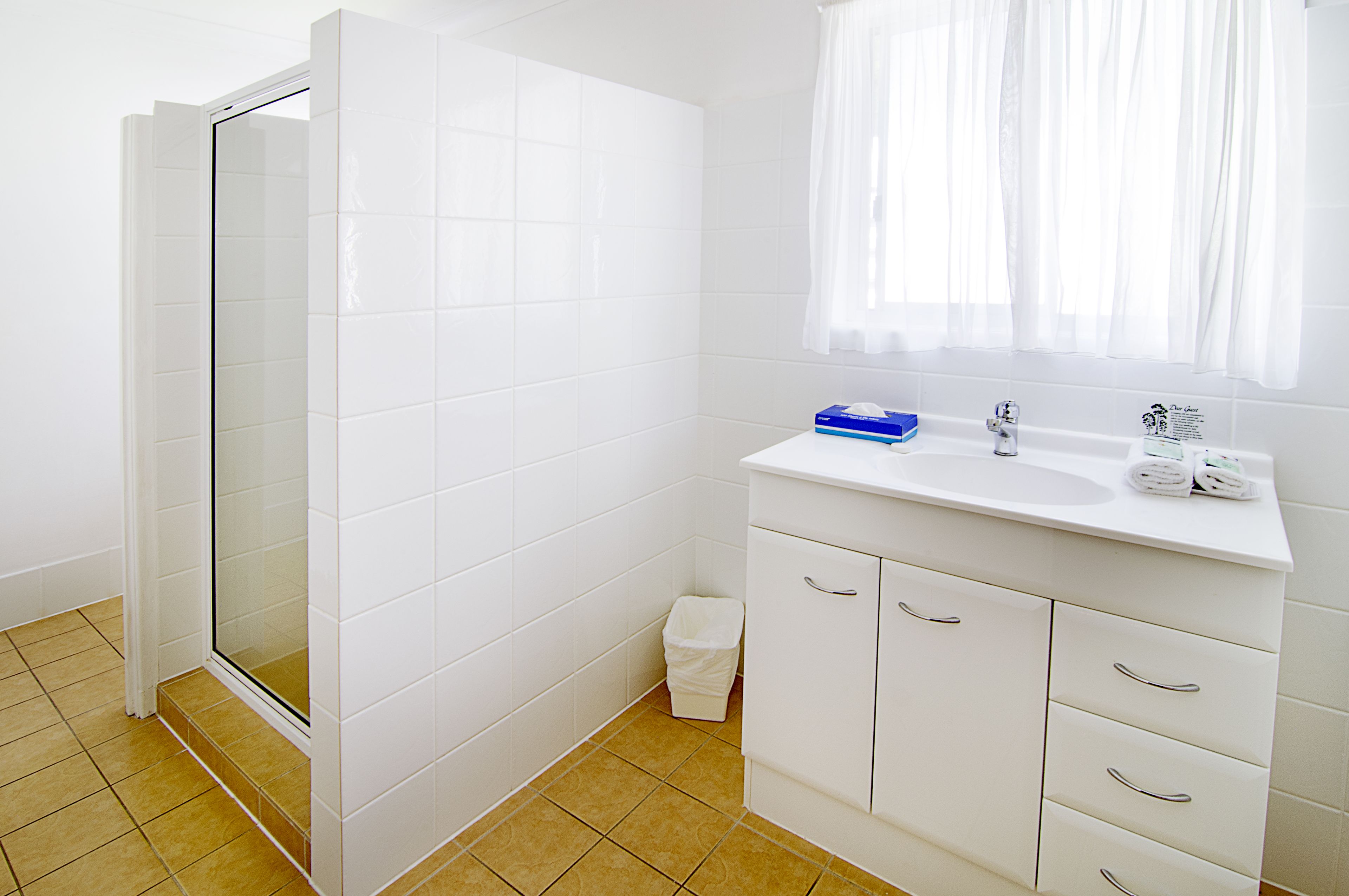 A bathroom with white tiles, a shower, a sink, and a window with curtains.