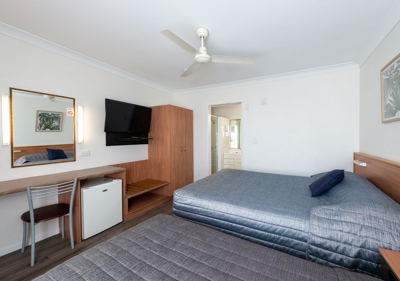 Ein kleines Hotelzimmer mit einem Bett, einem Schreibtisch, einem Stuhl, einem kleinen Kühlschrank, einem Deckenventilator und einem Fernseher an der Wand.