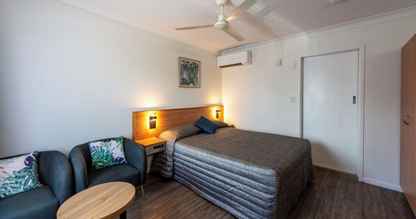 A bedroom with a bed, a nightstand, a chair, a table, a ceiling fan, and an air conditioning unit.