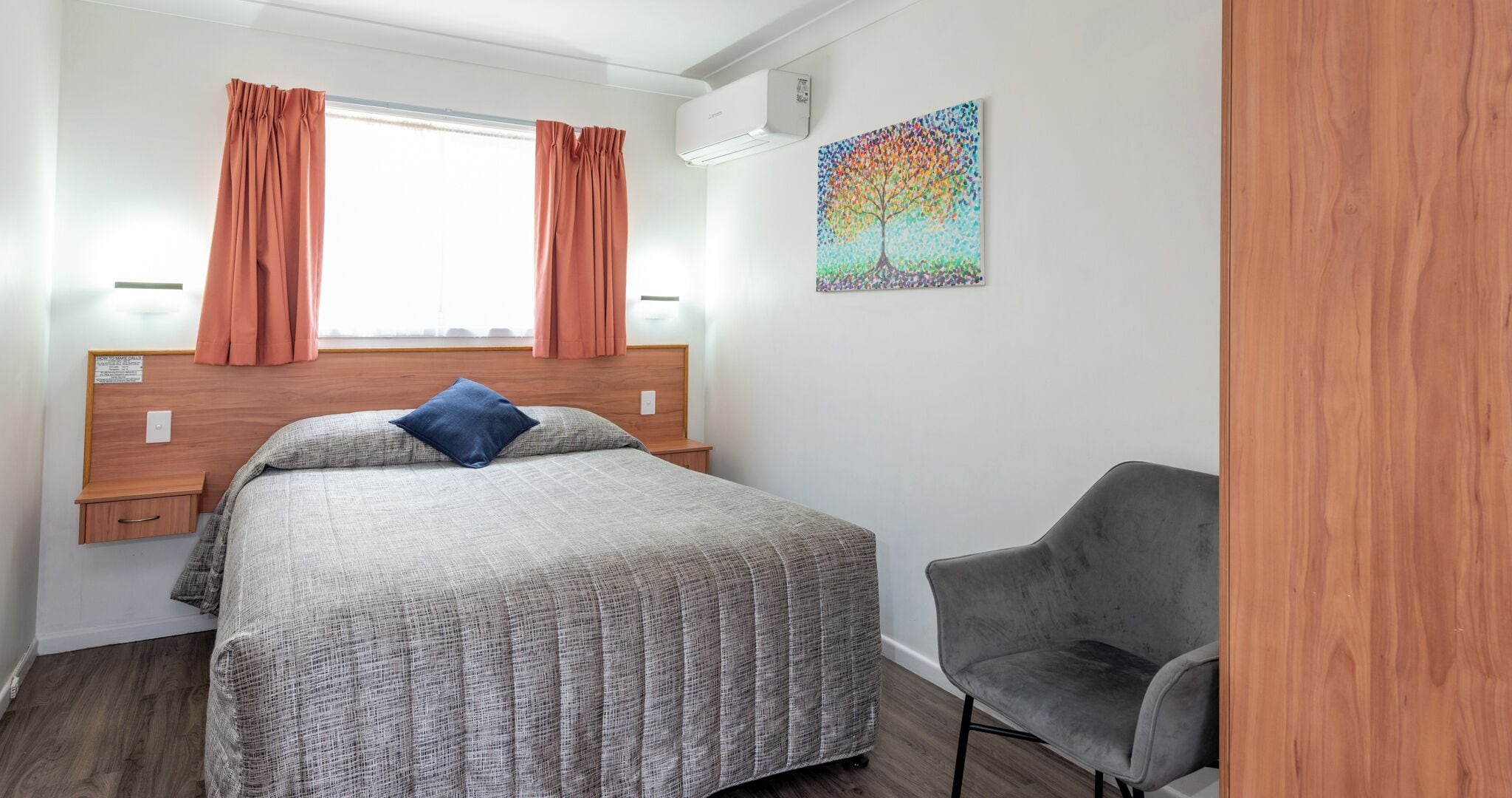 A hotel room with a bed, a chair, a window with curtains, a wall-mounted air conditioner, and a colorful artwork.