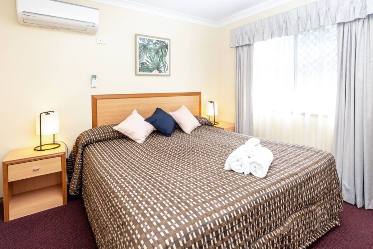 Una camera d'albergo con un letto grande, un comodino, un condizionatore d'aria, un quadro appeso alla parete e una finestra con tende.