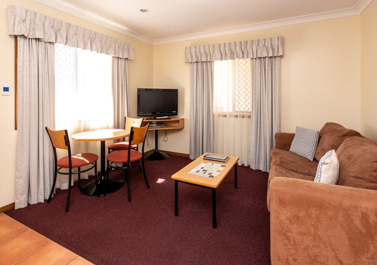 A hotel room with a brown sofa, a wooden coffee table, a TV, a dining table with chairs, and light-colored curtains.