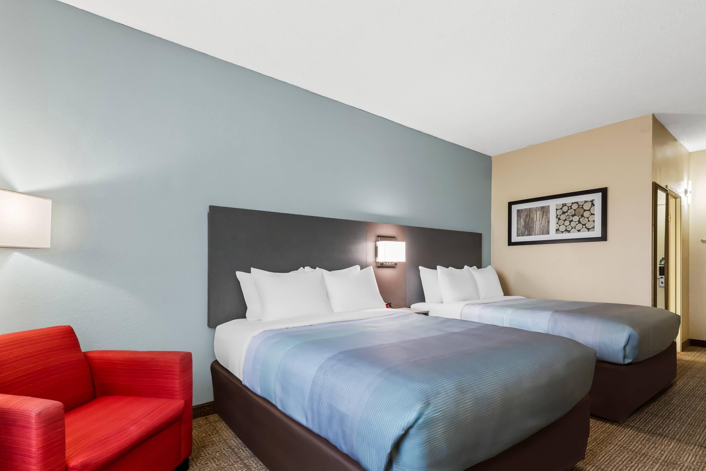 Une chambre d’hôtel avec deux lits, un fauteuil rouge et une lampe fixée au mur.
