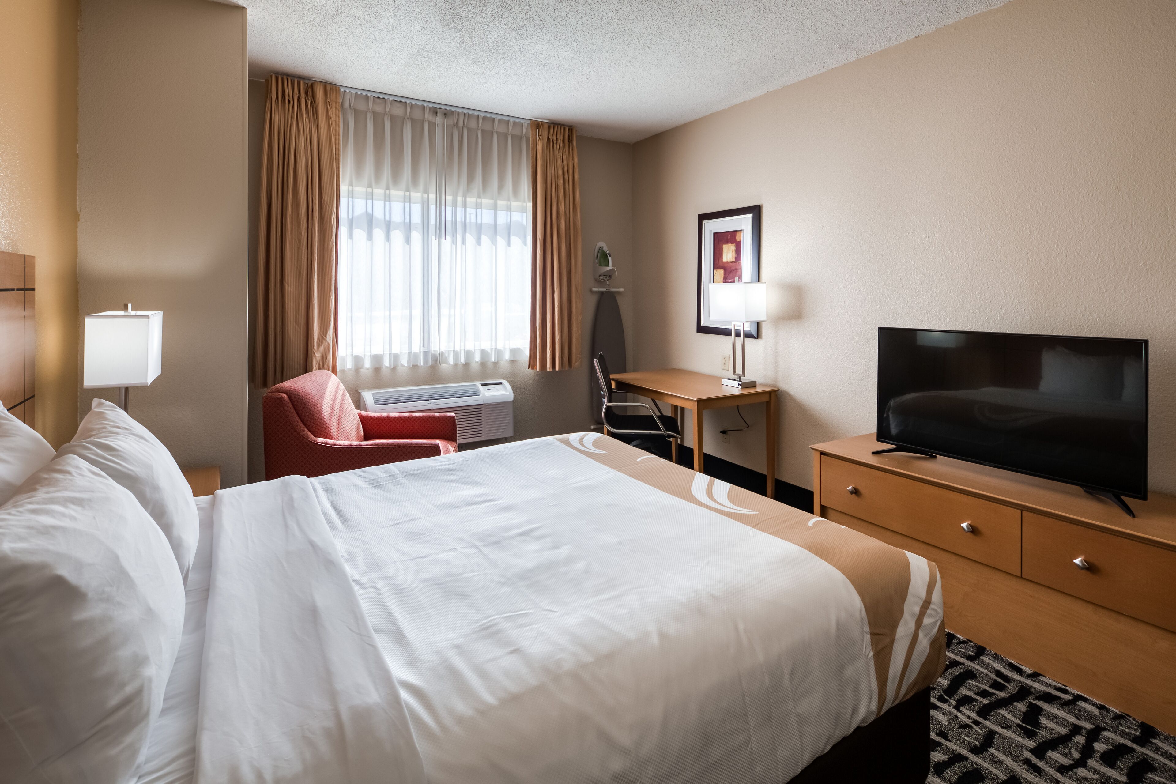 A hotel room with a large bed, a desk with a chair, a television, and a window with curtains.