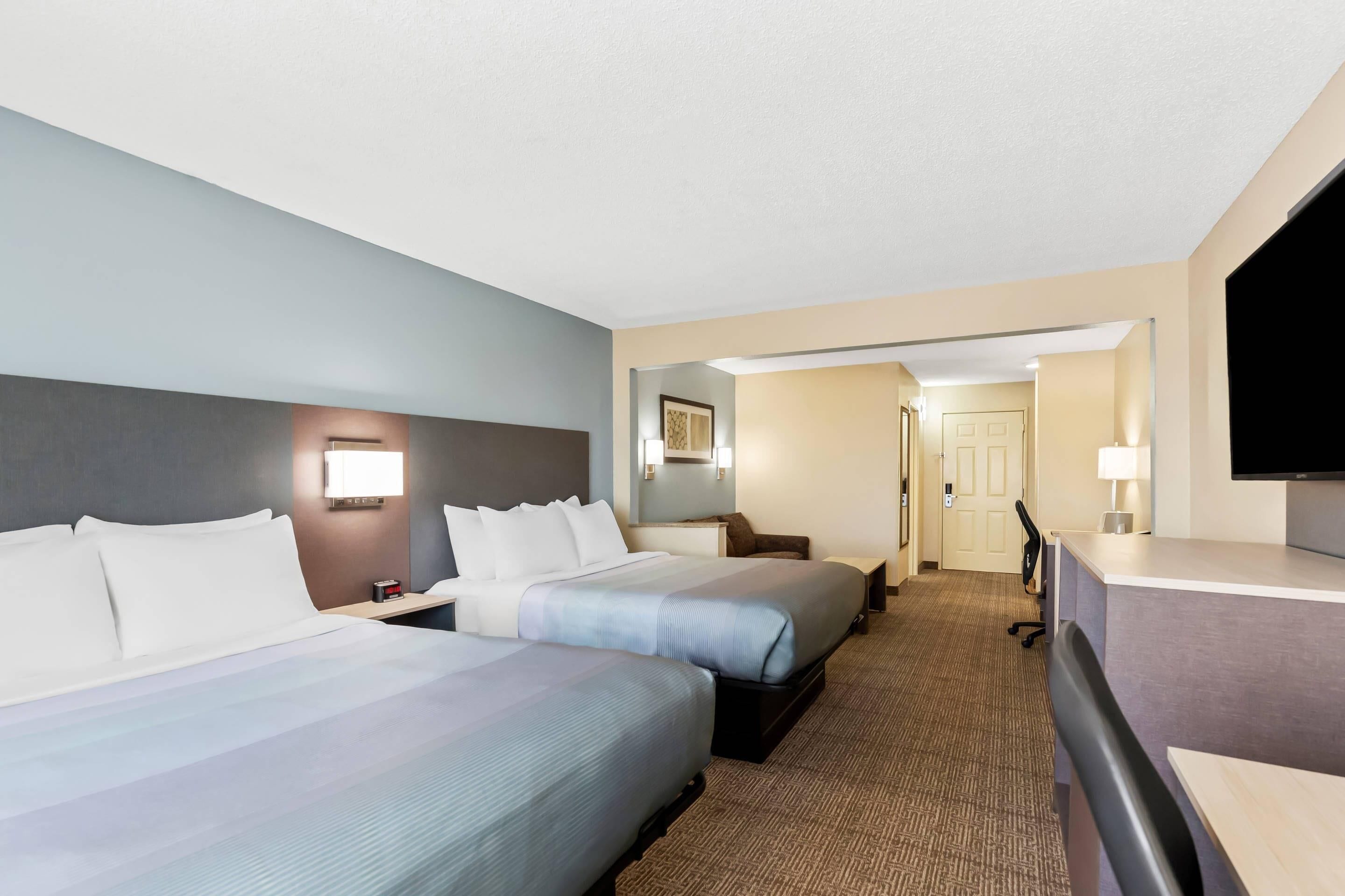 A hotel room with two beds, a desk, a TV, and a door leading to a hallway.