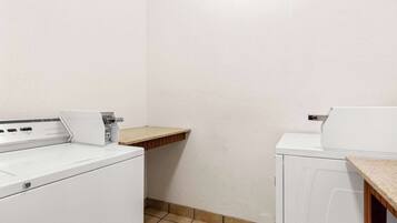 A laundry area with two white washing machines, a wooden shelf, and a tiled floor.