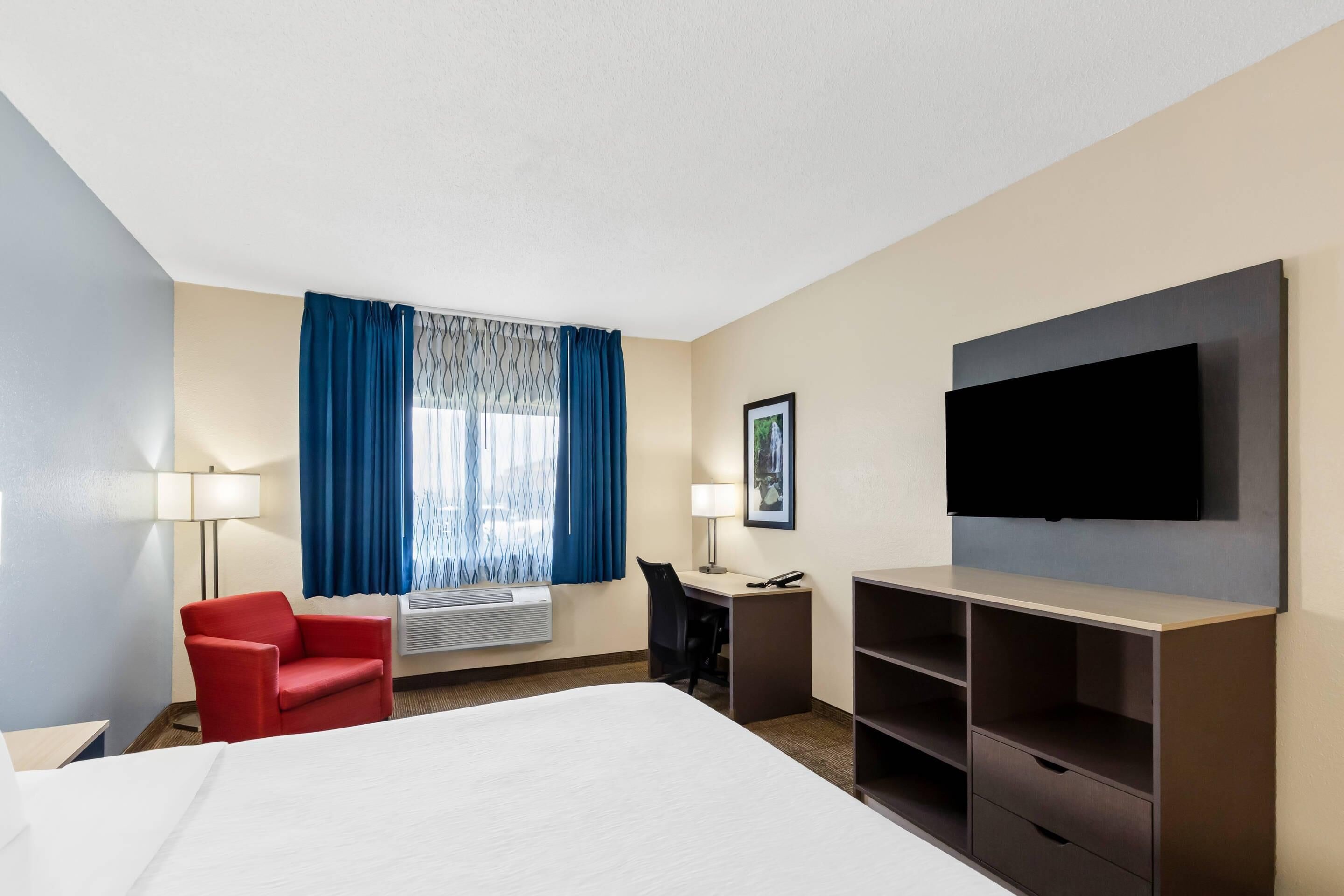 Quarto de hotel com cama, escrivaninha, cadeira, televisão e janela com cortinas.